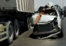 White sedan severely damaged after colliding with a large truck trailer on a highway debris and damaged bumper in the road responders in the background