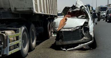 White sedan severely damaged after colliding with a large truck trailer on a highway debris and damaged bumper in the road responders in the background