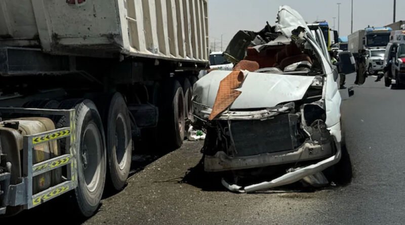 White sedan severely damaged after colliding with a large truck trailer on a highway; debris and damaged bumper in the road, responders in the background.